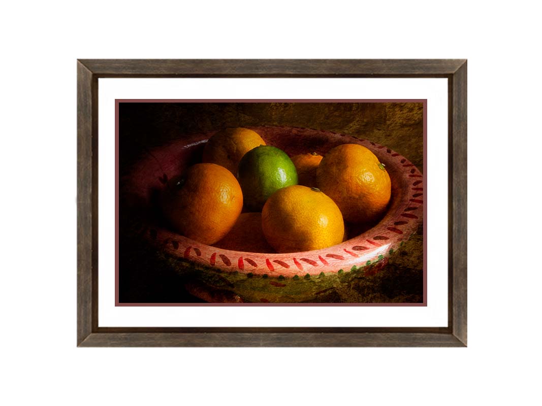 A photo of a bowl of oranges, with one lime placed in the center. Printed on paper, matted, and framed.