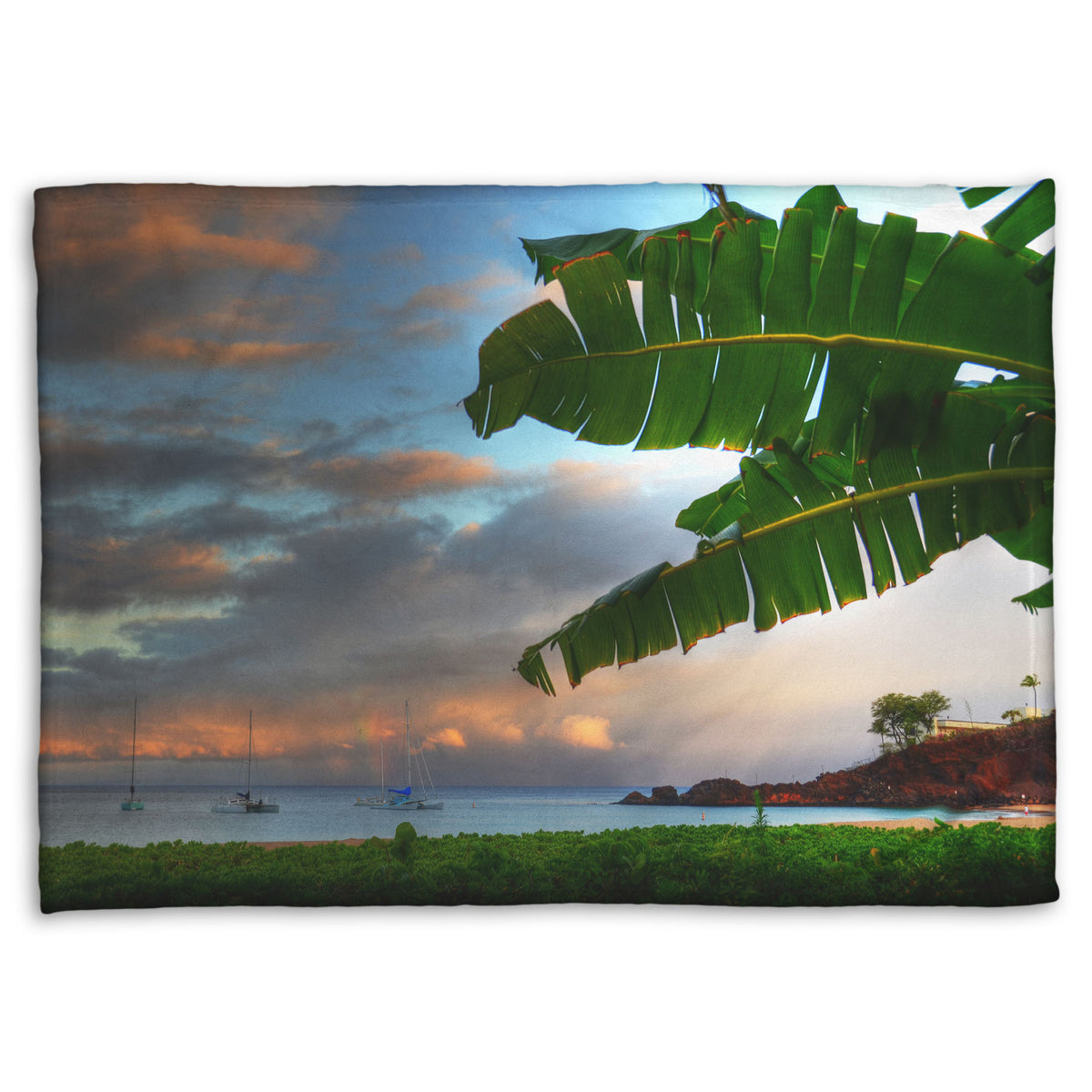 A fleece throw blanket designed with a photo of Ka'anapali beach in Hawai'i. Boats can be seen sailing on the ocean, and bright green plants grow in the foreground alongside a palm.