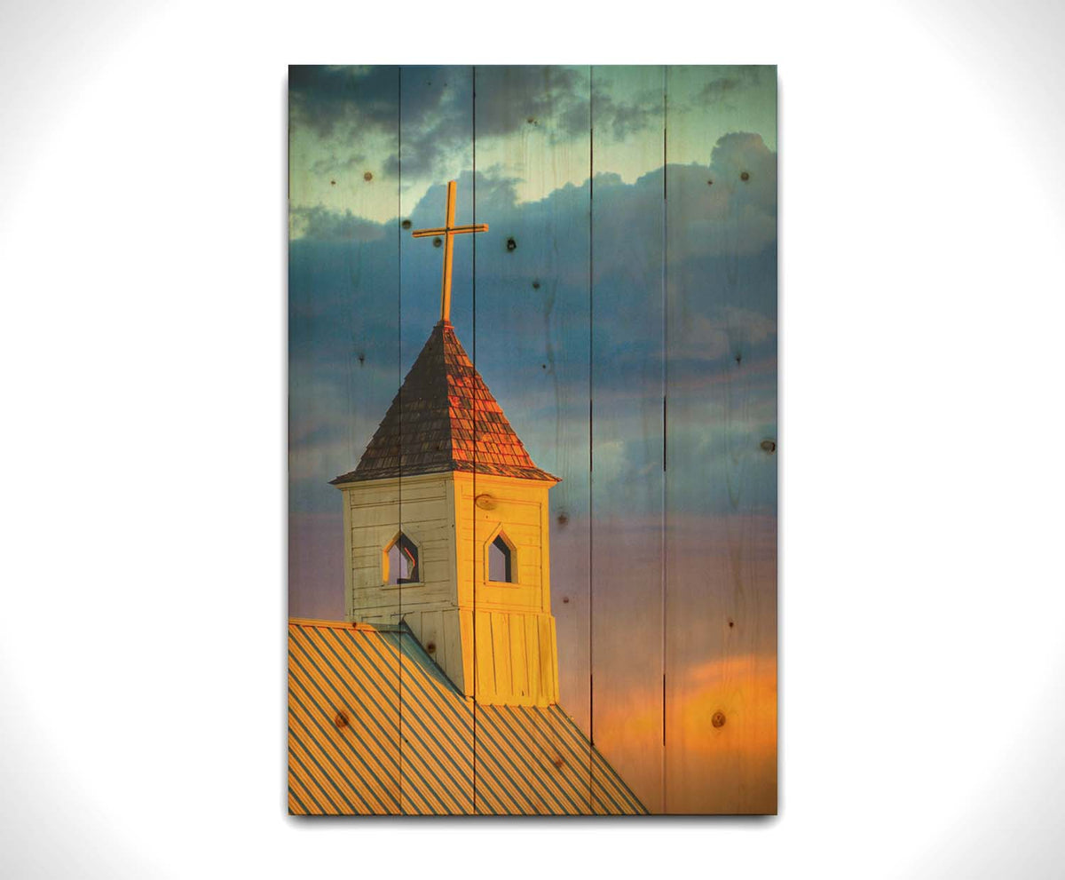 A photo of a cross-topped church at sunset, the sky gradating from blue to purple to orange. Printed on a wood pallet.