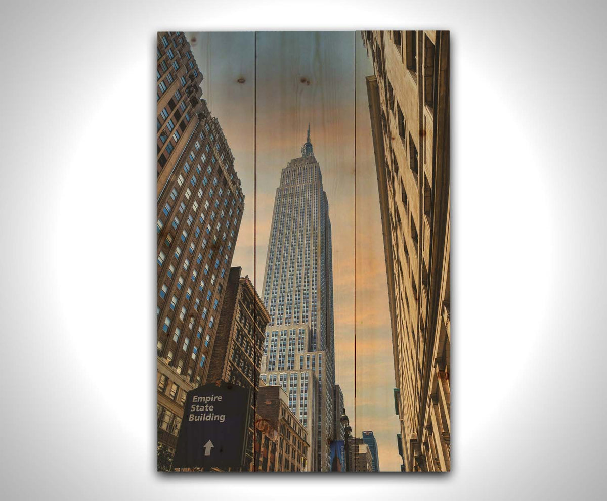 A photograph looking up at the Empire State Building in New York against a blue sky and orange clouds. Printed on a wood pallet.