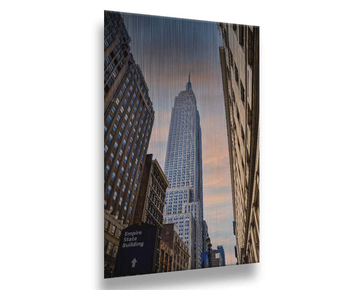 A photograph looking up at the Empire State Building in New York against a blue sky and orange clouds. Printed on metal.