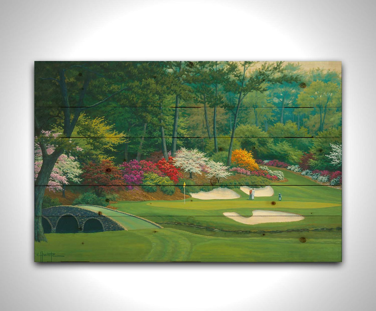 A painting of the Augusta Nationals golf course in Georgia. The course is encircled by foliage and flowers of all colors, and a bridged stream runs through the center. A few golfers stand near a small sand pit, lining up a shot to the nearby hole. Printed on a wood pallet.
