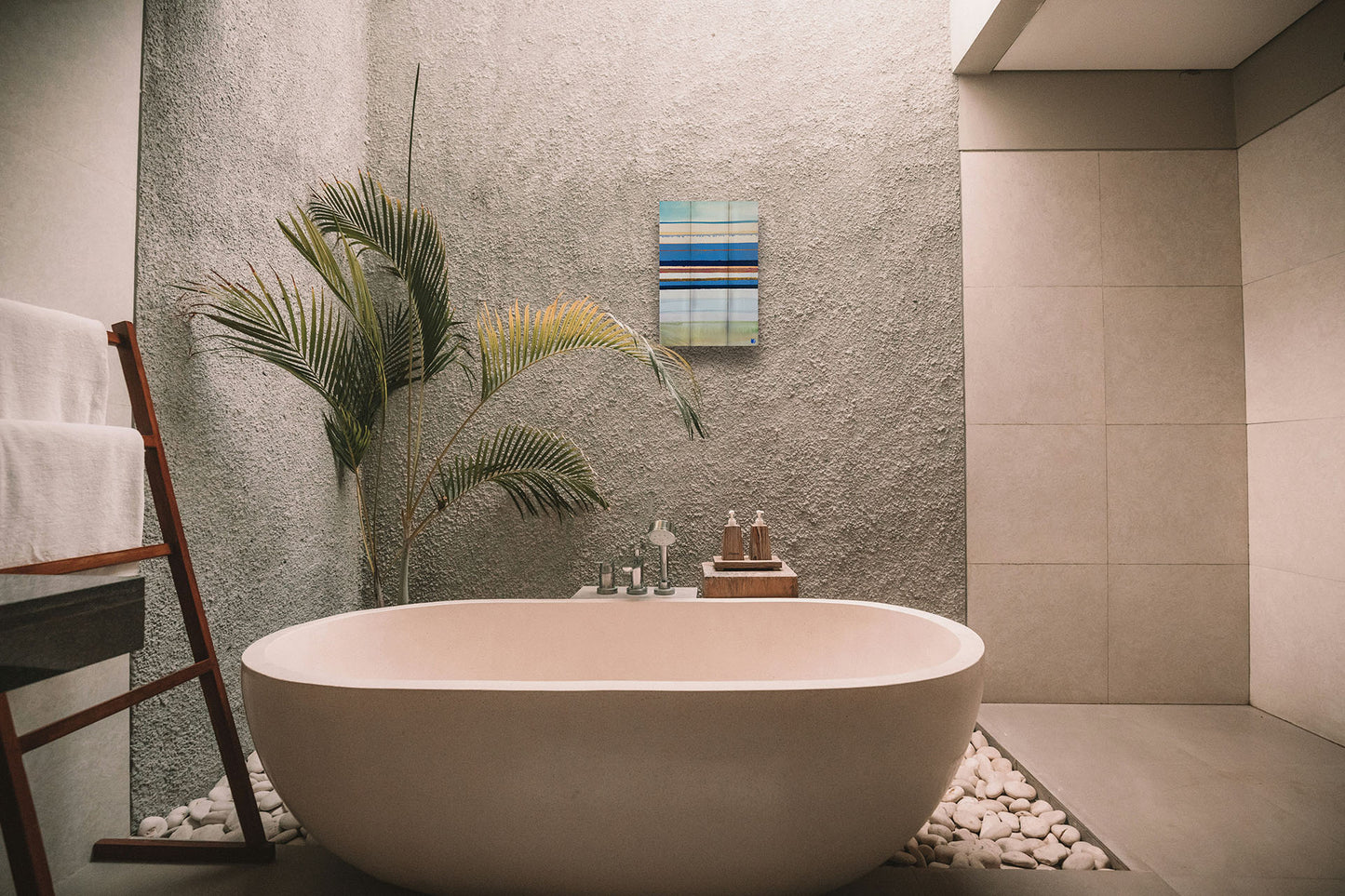 A painting hanging on a bathroom wall. An abstract painting arranging blue, white, yellow, and gold horizontal lines. The arrangement suggests the feeling of a shoreline. Printed on a box board.