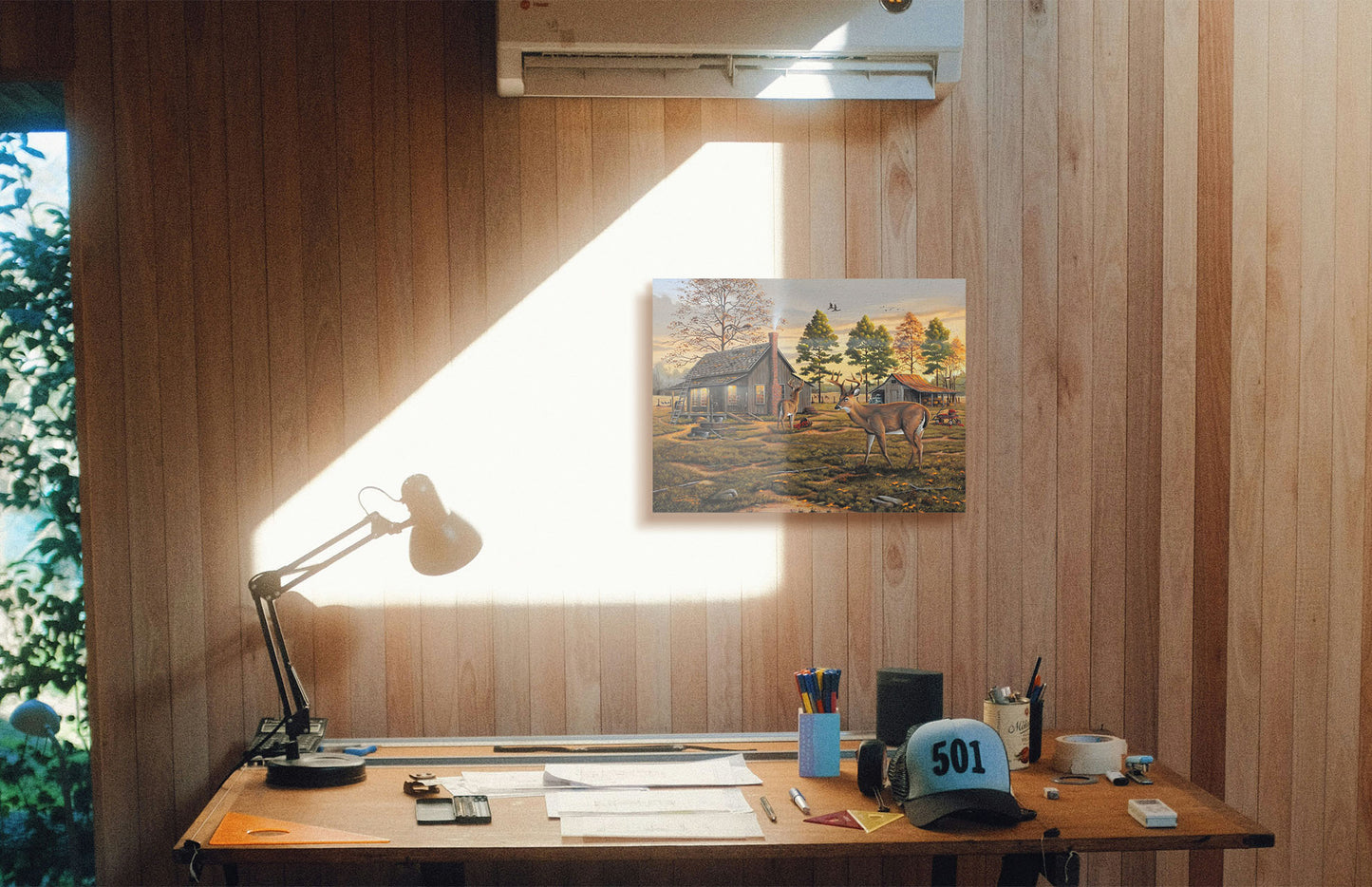 A painting hanging on a study wall. A painting of a farmhouse scene, featuring a fenced field, wood cabin, and an accompanying shed. In the foreground, deer are eating apples from wagons and baskets in the yard. Printed on canvas.