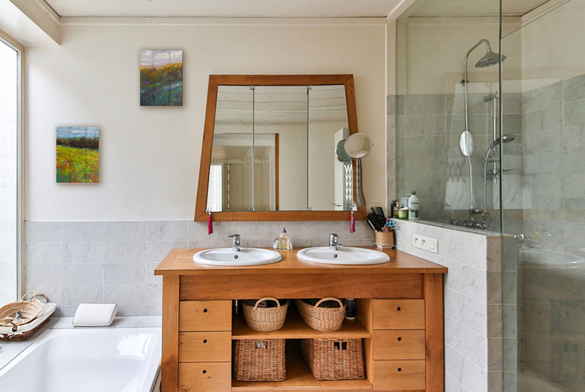 Two paintings hanging on a bathroom wall. The first is a landscape painting of a colorful meadow bordered by a lush forest, created using vibrant green impasto with pops of orange, pink, yellow, and blue. Printed on a box board. The second is a painting of a grassy highland hillside, created with visible brushstrokes. Printed on a box board.