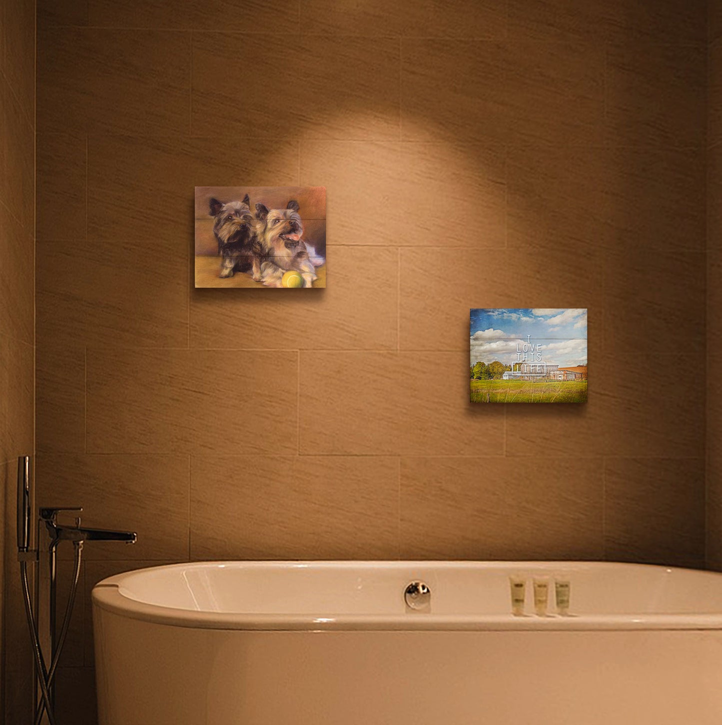 A painting and a photograph hanging on a bathroom wall. The first is a painting of two brown terrier dogs sitting with a tennis ball. Printed on a box board. The second is a photo of several barns, their associated pastures, and fields. The early signs of fall are present in the steadily changing leaves and crops. The sky above is wide, blue, and populated with fluffy clouds. It is overlaid with the phrase "I love this life." Printed on a box board.