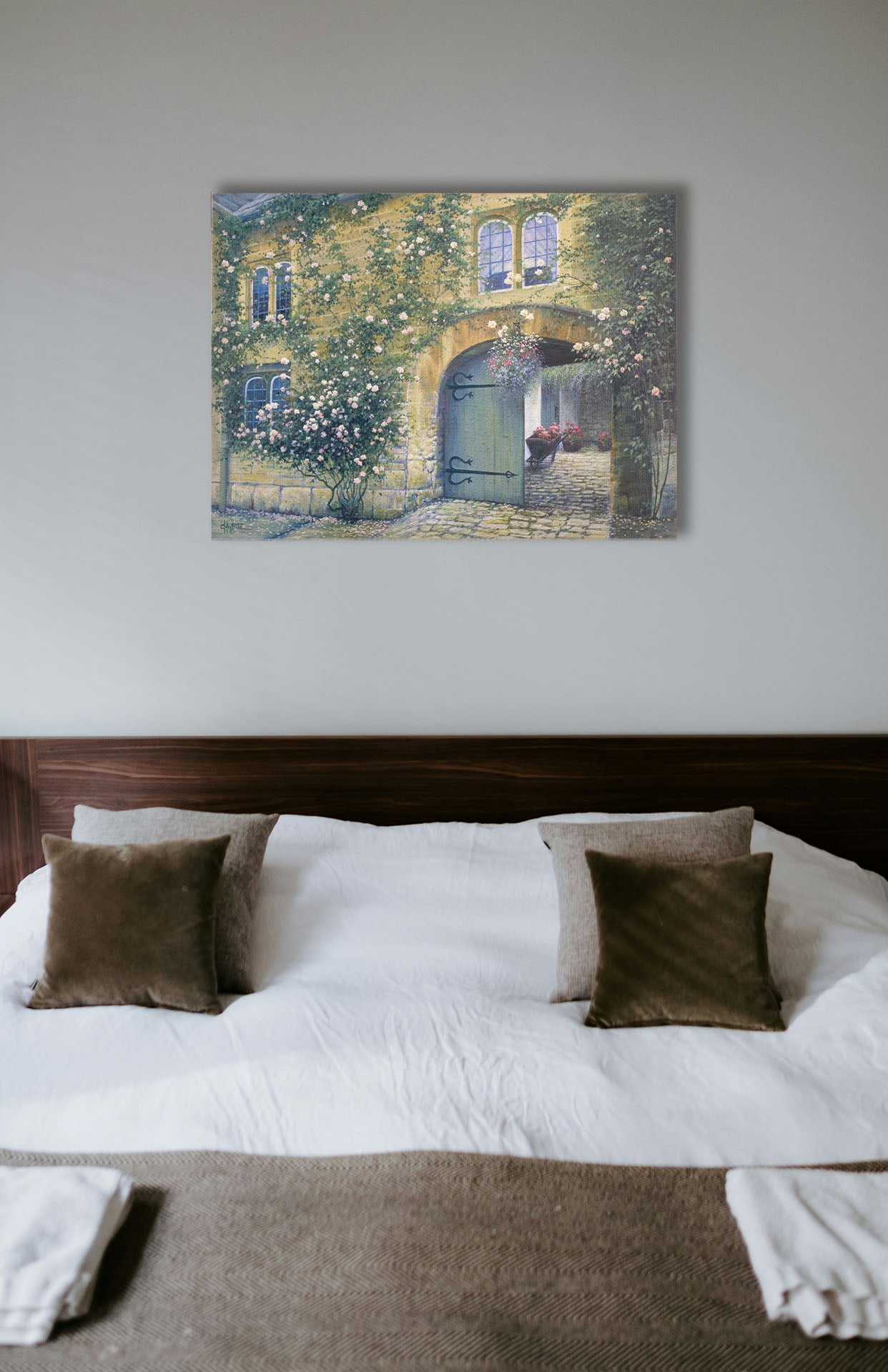 A painting hanging on a bedroom wall. A painting of an arched doorway passing through a building into a courtyard. The building and courtyard are decorated with bushes and vines sprouting an assortment of white and red flowers. Printed on canvas.