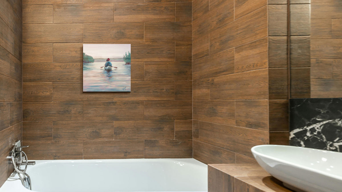 A painting hanging on a bathroom wall. A painting of two people paddling a canoe across a lake in the early morning. The sky is pale reflecting on the water, and green trees line the shore along the lake. Printed on a box board.