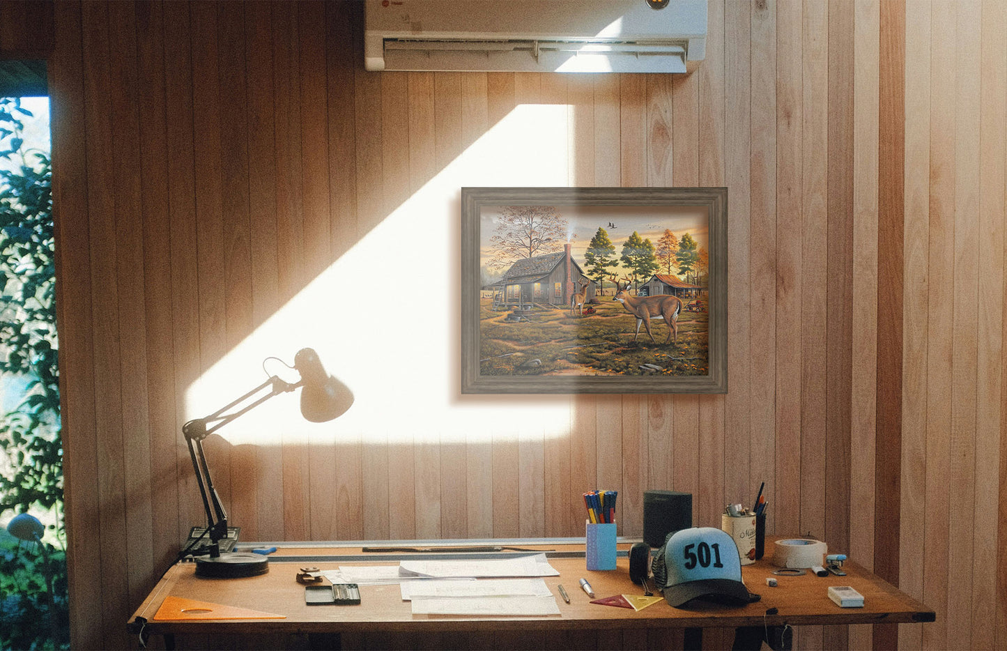 A painting hanging on a study wall. A painting of a farmhouse scene, featuring a fenced field, wood cabin, and an accompanying shed. In the foreground, deer are eating apples from wagons and baskets in the yard. Printed on canvas and framed.