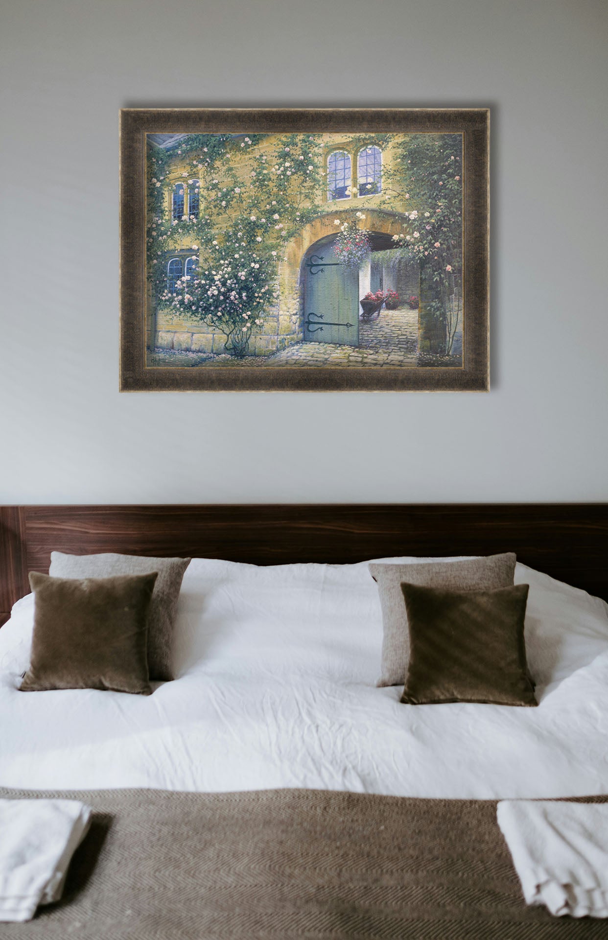A painting hanging on a bedroom wall. A painting of an arched doorway passing through a building into a courtyard. The building and courtyard are decorated with bushes and vines sprouting an assortment of white and red flowers. Printed on canvas and framed.