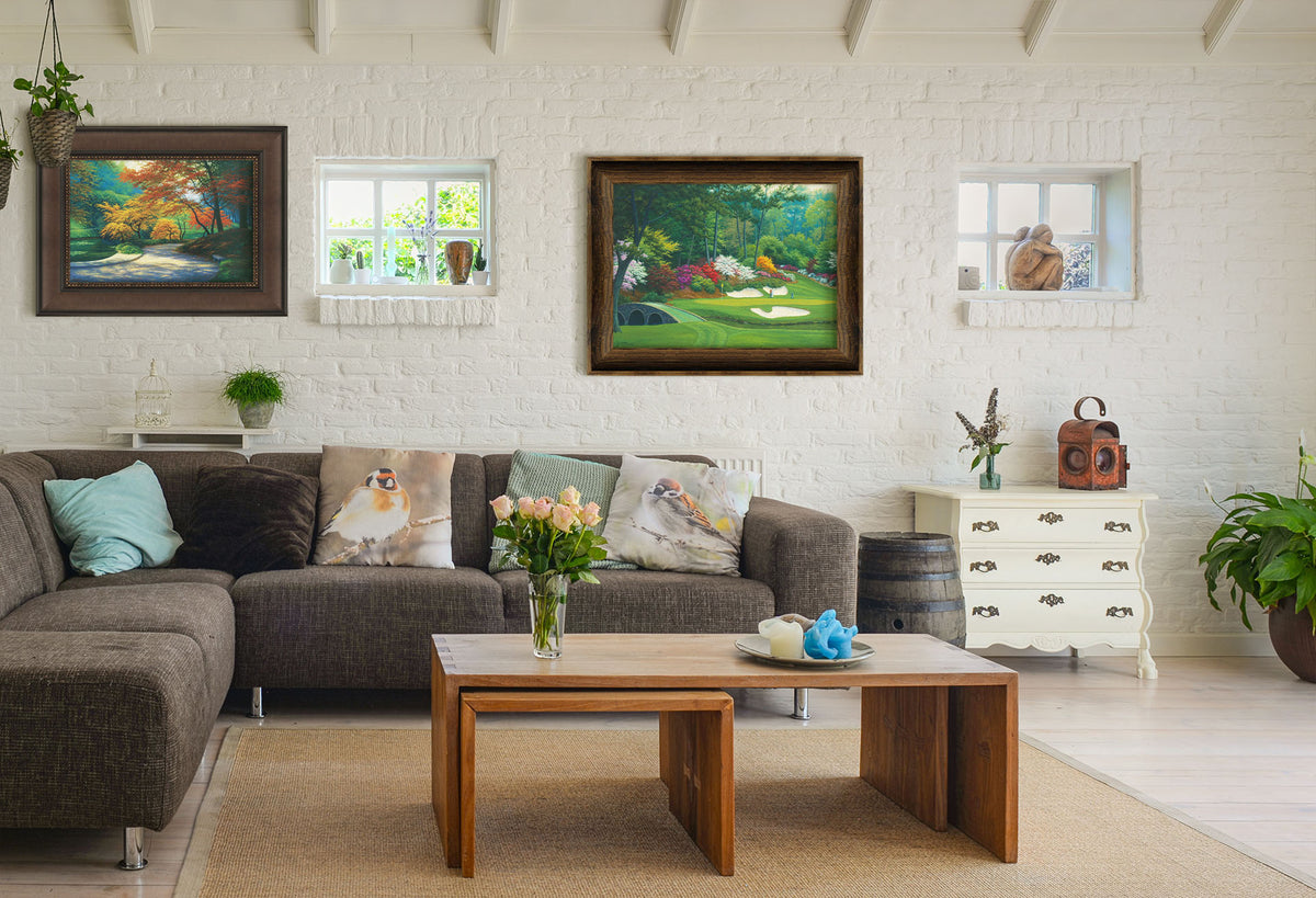Two paintings hanging on a living room wall. The first is a painting of a paved garden path beside a pond, bordered by orange and yellow fall trees. Printed on canvas and framed. The second is a painting of the Augusta Nationals golf course in Georgia. The course is encircled by foliage and flowers of all colors, and a bridged stream runs through the center. A few golfers stand near a small sand pit, lining up a shot to the nearby hole. Printed on canvas and framed.