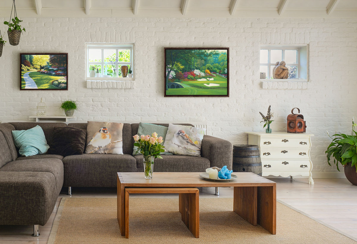 A painting hanging on a living room wall. The first is a painting of a cobblestone path through a sunny, colorful, tidy garden. Printed on canvas in a float frame. The second is a painting of the Augusta Nationals golf course in Georgia. The course is encircled by foliage and flowers of all colors, and a bridged stream runs through the center. A few golfers stand near a small sand pit, lining up a shot to the nearby hole. Printed on canvas in a float frame.