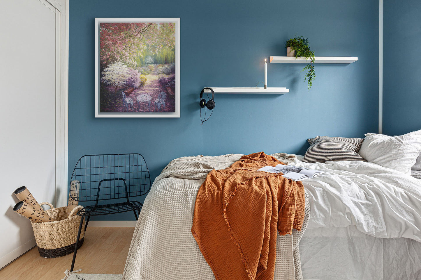 A painting hanging on a bedroom wall. A painting of a springtime garden, with cherry blossom petals coating the garden and pathways. White chairs, a table, and a bench decorate the garden full of red, pink, and white flowers. Printed on canvas in a float frame.