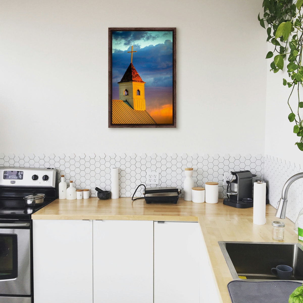 A photograph hanging on a kitchen wall. A photo of a cross-topped church at sunset, the sky gradating from blue to purple to orange. Printed on canvas in a float frame.