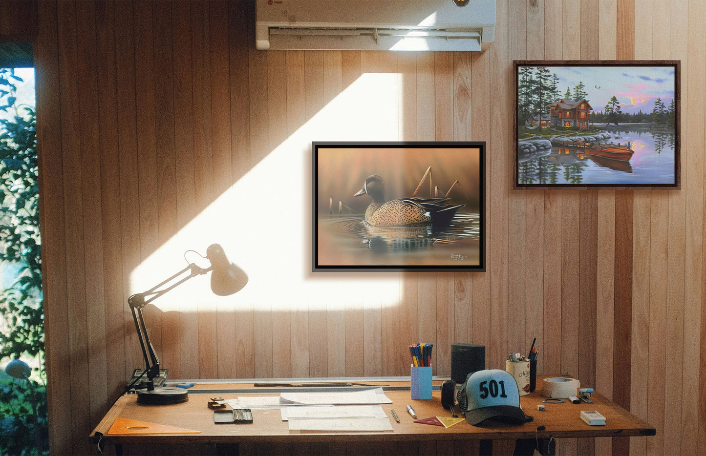 Two paintings hanging on a study wall. The first is a painting of a blue-winged teal waterfowl, swimming across the water past several brown reeds. Printed on canvas in a float frame. The second is a painting of a lakeside cabin, with a boat out on the water and geese overhead. Printed on canvas in a float frame.