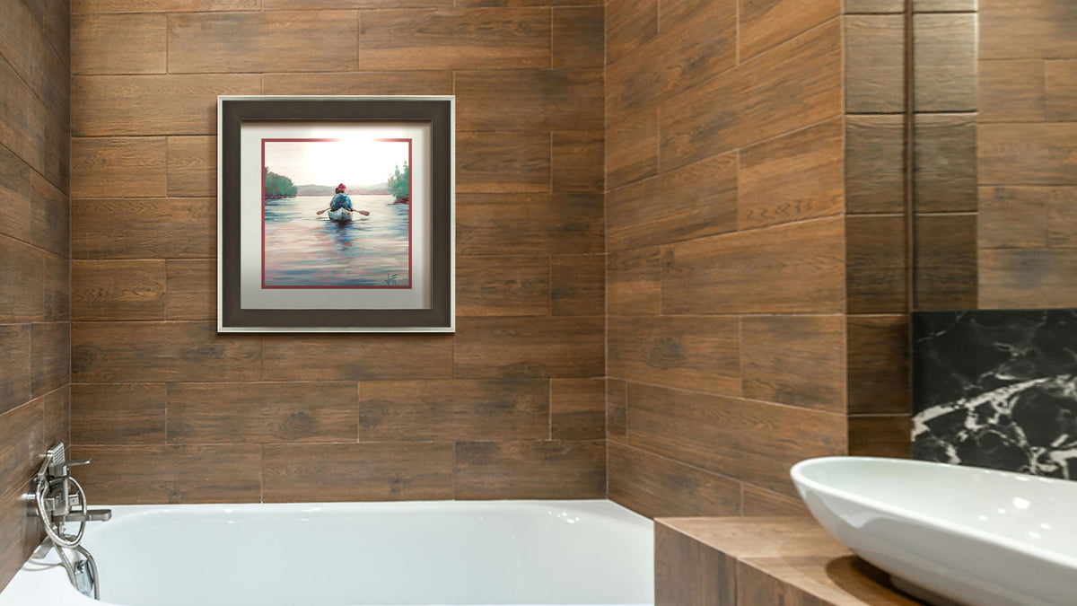 A painting hanging on a bathroom wall. A painting of two people paddling a canoe across a lake in the early morning. The sky is pale reflecting on the water, and green trees line the shore along the lake. Printed on paper, matted, and framed.