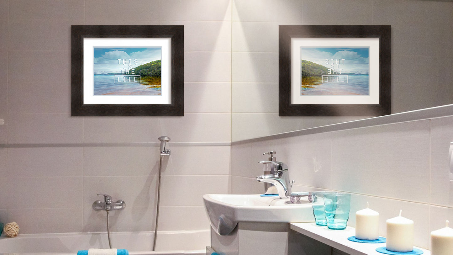 A photograph hanging on a bathroom wall. A photograph taken from the shore, looking out toward a forested coast. The water reflects the blue sky and gentle clouds. The phrase "This is the life" is overlaid across the center of the photo in white. Printed on paper, matted, and framed.