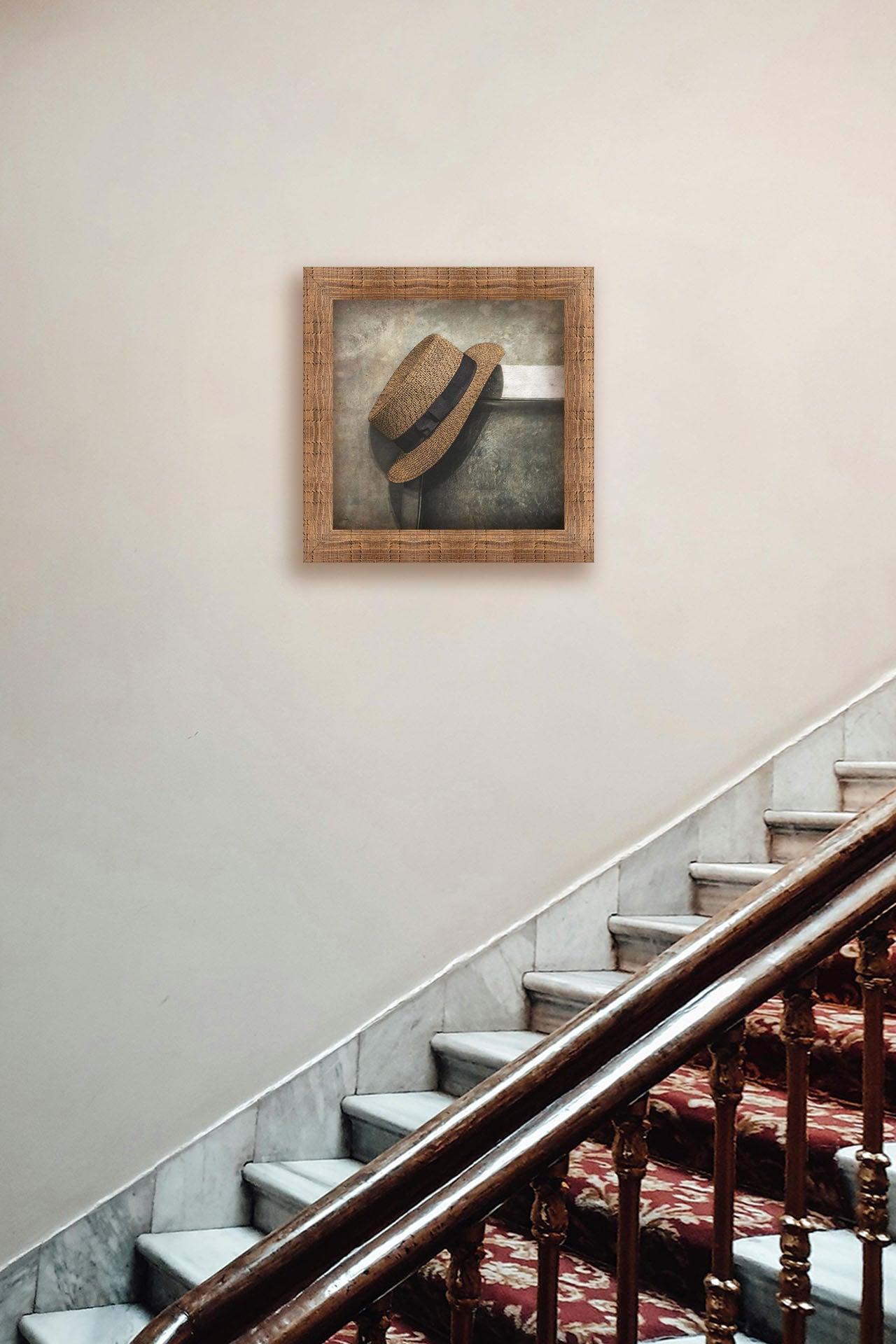 A photograph hanging on a hallway wall. A photograph of a brown boater hat hanging on the back of a chair, against a gray cement wall. Printed on canvas and framed.