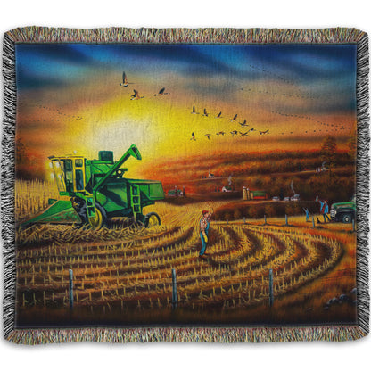 Woven blanket with a scenic design of a farm landscape, with a harvester stopped in a partially harvested field of corn.