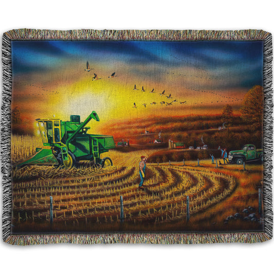 Woven blanket with a scenic design of a farm landscape, with a harvester stopped in a partially harvested field of corn.