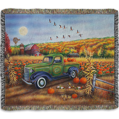 A woven throw blanket designed with a painting of a farm, featuring a field of pumpkins and a field of corn. There is a forest of yellow, orange, red, and green fall trees in the background, and geese fly overhead.