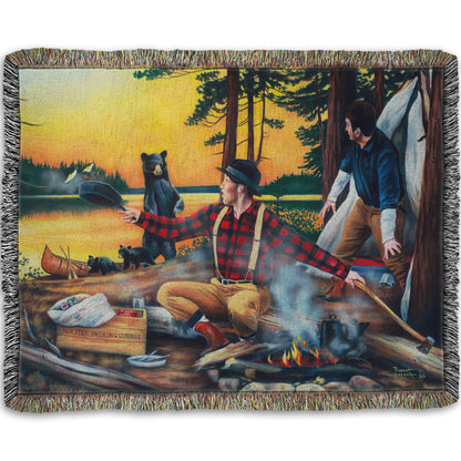 Woven throw blanket designed with a painting of two men camping and cooking a meal, looking back in shock at an approaching bear and her two cubs.