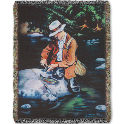 A woven throw blanket designed with a painting of a fisherman standing in the river, cleaning a fish he just caught.