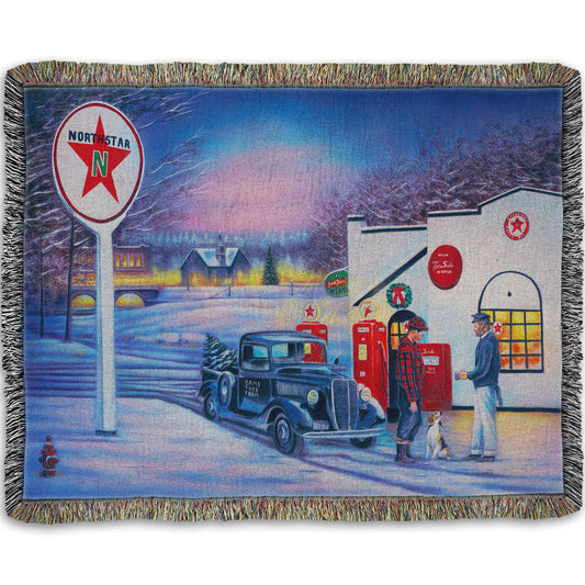 A woven throw blanket decorated with a 1950s Americana scene, with a vintage truck refueling at a Northstar gas station.