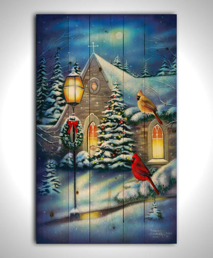 A painting of cardinals perched on evergreen branches on a snowy night, in front of a church decorated for Christmas. Printed on a wood pallet.