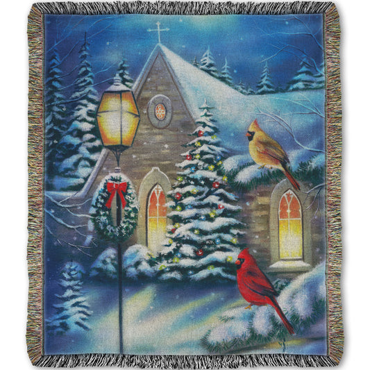 Woven throw blanket designed with a snowy christmas scene in a church courtyard, where cardinals are perched on evergreen tree branches.