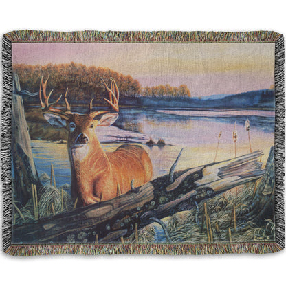 Woven throw blanket designed with a painting of a deer in front of a lake reflecting the morning sky.