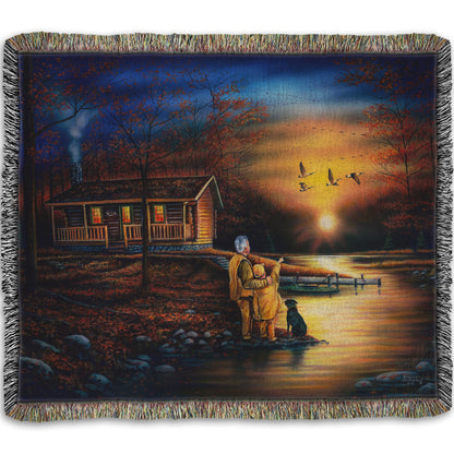 A woven throw blanket designed with a painting of a man and a child standing by a river and cabin. They are watching geese fly overhead at sunset.