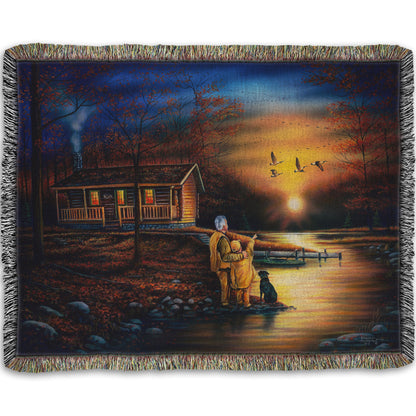 A woven throw blanket designed with a painting of a man and a child standing by a river and cabin. They are watching geese fly overhead at sunset.