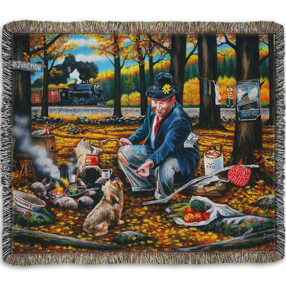 A woven throw blanket designed after a painting of a man sitting on a log at a campfire in the woods strewn with fall leaves. He is feeding a little dog. A train is passing in the background.