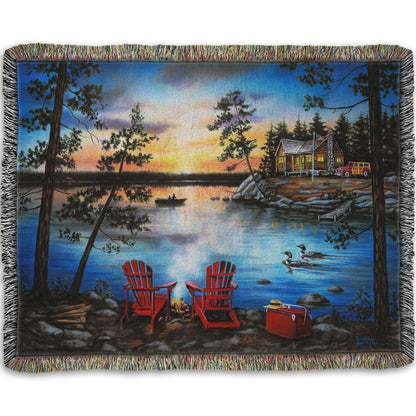 A woven throw blanket designed after a painting of a lake at sunset, with a campfire in the foreground. Two loons drift across the water, as well as two fishers.