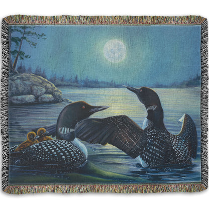 A woven throw blanket designed with a painting of two grown loons, with two baby loons on one of their backs. They are floating on a lake under the moonlight.
