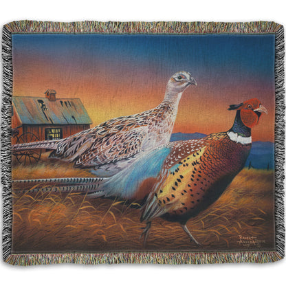Woven throw blanket designed with a painting of two pheasants, walking through golden grasses against a colorful sunset.