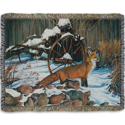 A woven blanket designed with a painting of a red fox in the winter snow. It is standing by some old farming equipment, grasses, and stones.