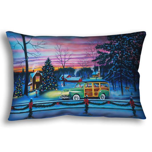A throw pillow printed with a painting of a snowy town with Christmas lights, where a 1950s family drive past in a car full of presents at sunset.