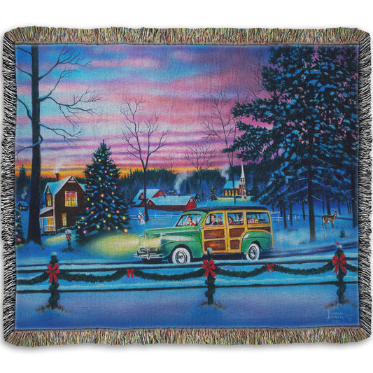 A woven thrown blanket printed with a painting of a snowy town with Christmas lights, where a 1950s family drive past in a car full of presents at sunset.