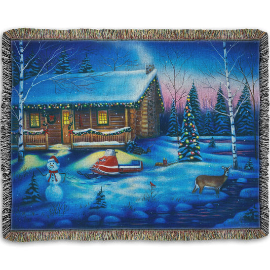 A festive woven throw blanket decorated with the painting of a snowy cabin decorated with Christmas lights.