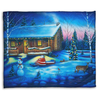 A festive coral fleece blanket decorated with the painting of a snowy cabin decorated with Christmas lights.