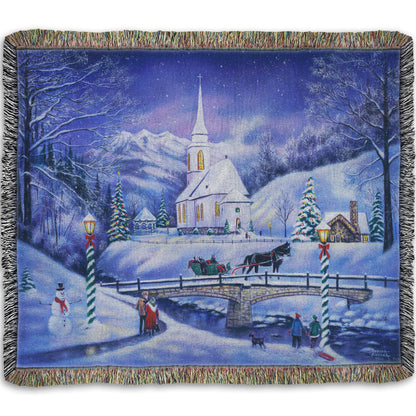 A woven throw blanket designed with a snowy mountain town, decorated for Christmas. A tall church sits in the middle of town.