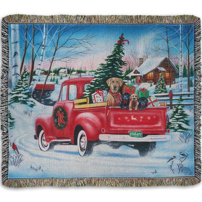 Festive woven blanket with a red truck carrying a Christmas tree, puppies, and presents in a snowy village scene.