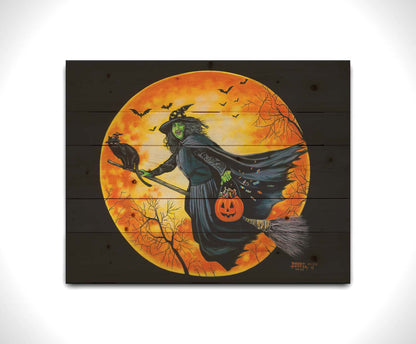 A painting of a witch, flying on a broom in front of an orange moon while carrying a pumpkin-shaped basket of candy. Printed on a wood pallet.