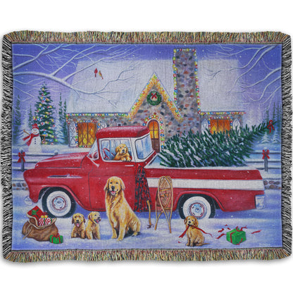 A throw blanket woven to design a painting of a vintage red truck carrying a Christmas tree to a decorated house, surrounded by a family of golden retriever dogs.