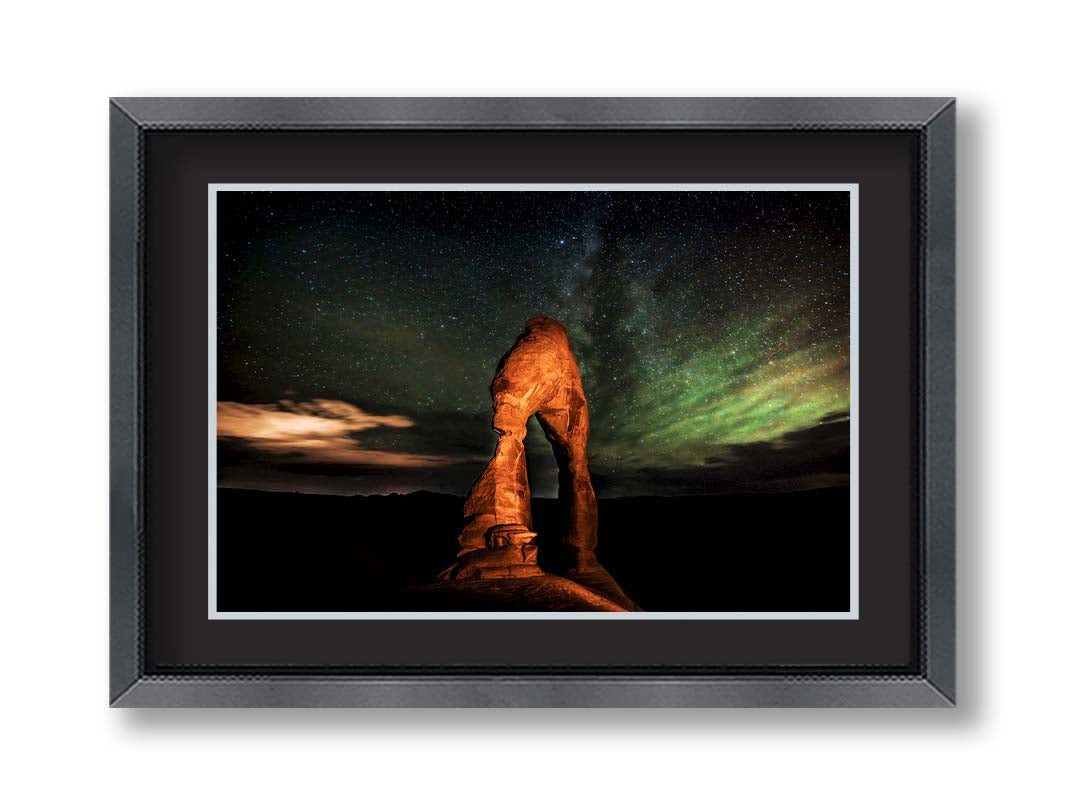 Nighttime photo of Delicate Arch in Arches National Park, Utah. The arch is lit with staged lighting, with a backdrop of a starry night sky. Printed on paper, matted, and framed.