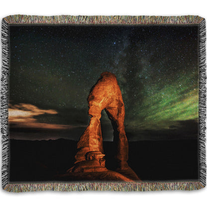 Woven throw blanket designed with a nighttime photo of Delicate Arch in Arches National Park, Utah. The arch is lit with staged lighting, with a backdrop of a starry night sky.