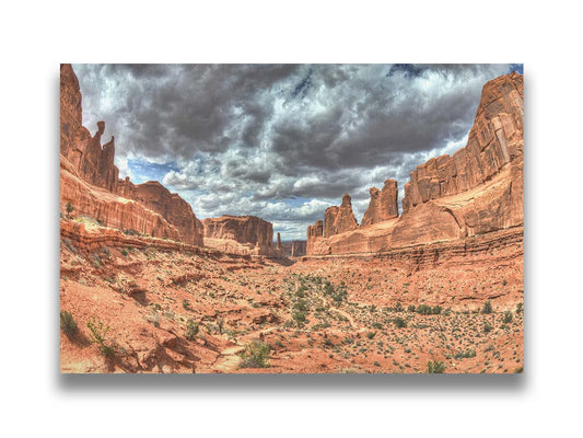 A photo of a tan, sandy, rocky valley along the Park Avenue hiking trail at Arches National Park in Utah. Gray and white clouds fill the sky. Printed on canvas.