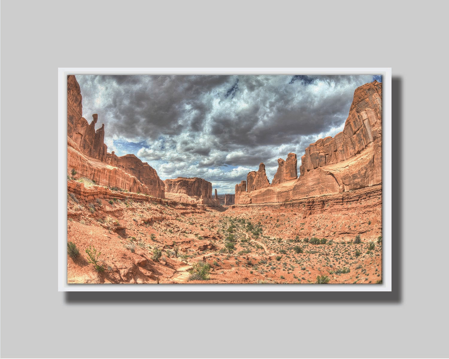 A photo of a tan, sandy, rocky valley along the Park Avenue hiking trail at Arches National Park in Utah. Gray and white clouds fill the sky. Printed on canvas in a float frame.